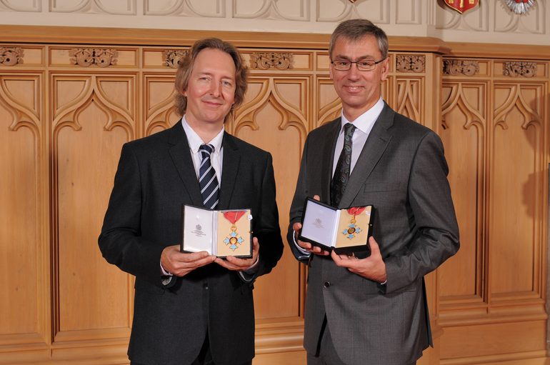 Jonathan Church on the left, and Alan Finch on the right, stand holding their CBEs in front of a wooden Gothic screen.
