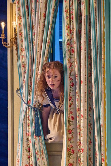 A girl sneaks through window. There are ornate, floral curtains and a warmly glowing sconce on the wall.