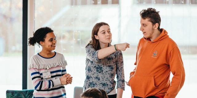 Three young people in a row standing and smiling. The girl in the middle is gesturing to the boy on her left, as if holding an imaginary microphone up to him.