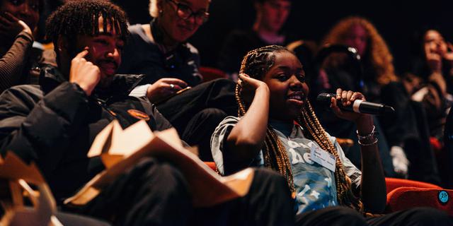 An audience of young people are seated in the Minerva theatre. A girl is speaking into the microphone.