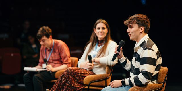 Three young people are seated on chairs on the Minerva stage. The boy in the foreground, wearing a black and white striped top, is speaking into a microphone.