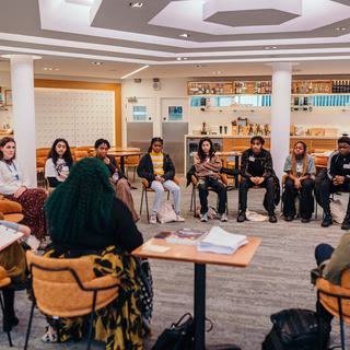 A large group of young people sit on orange chairs in a circle.