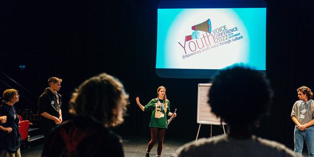 In a dark studio a group of young people are standing in a circle; behind them is a large blue screen with the Youth Conference logo on it.