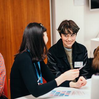 A boy in a red check shirt is seated to the left of the picture. Next to him, a girl in a black jumper with dark hair talks to the boy on her left.