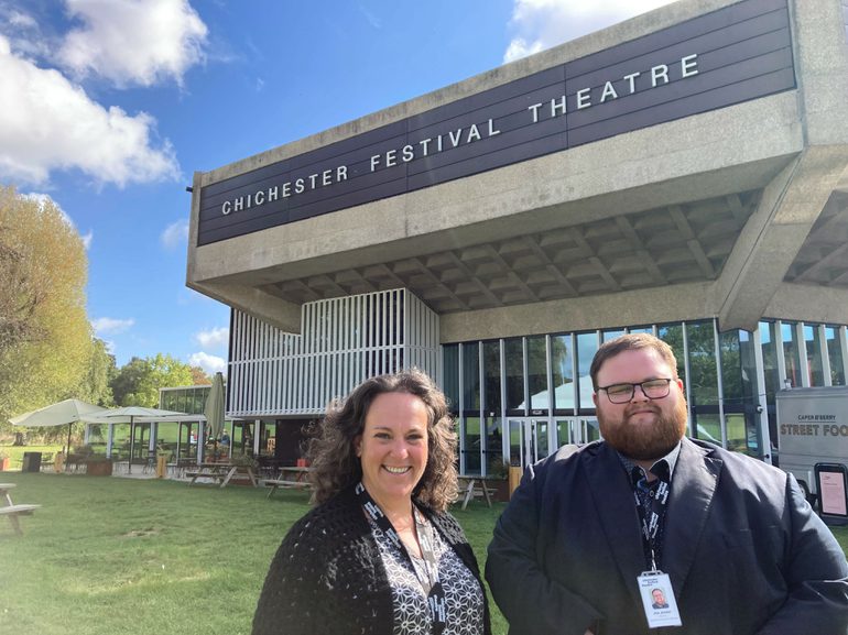 Sally and Joe stand smiling at the camera with Chichester Festival Theatre behind them. Sally is on the left with dark brown hair and waring a black and white shirt; Joe, on the right, has dark hair and a beard and is wearing black.