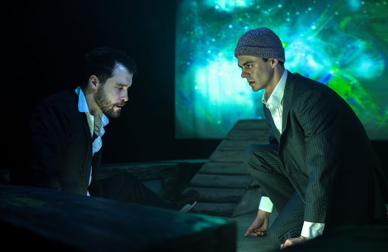Two young men sit facing each other with intense expressions. Behind them is a turquoise projection with a pattern like the sea or the night sky. They look tired, worried and worn down.