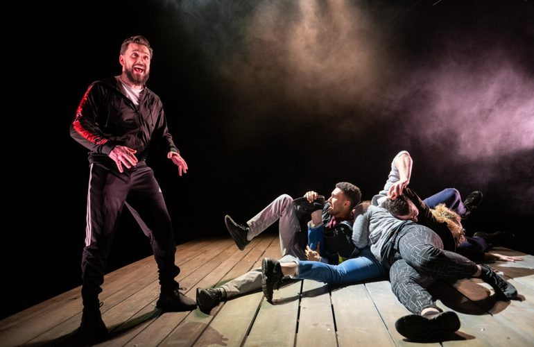 A man stands on a slanted wooden stage wearing a black tracksuit, speaking with fear or concern. On the ground, three others lie tangled together, in a pile.