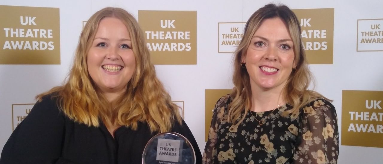 A woman in a black top and a woman in a flowery top stand in front of a banner displaying the UK Theatre Awards logo. They are smiling and holding an award.