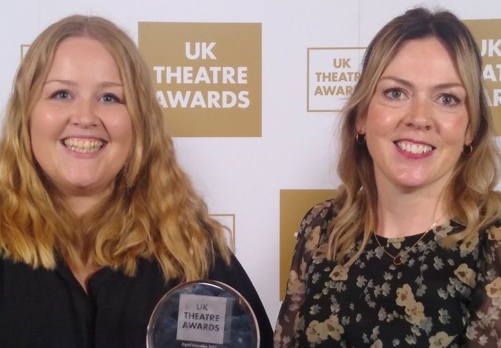 A woman in a black top and a woman in a flowery top stand in front of a banner displaying the UK Theatre Awards logo. They are smiling and holding an award.