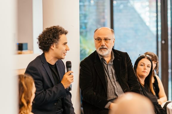 Two people sit talking at an event. One holds a microphone that he speaks into and the other looks at him with interest. They look comfortable together.
