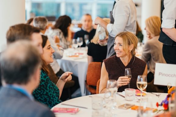 Two guests wearing smart clothes sit at a dinner table, drinking fizz. They smile and chat comfortably. Other guests can be seen around them and in the background and the table has been beautifully dressed with a white table cloth and colourful flowers.