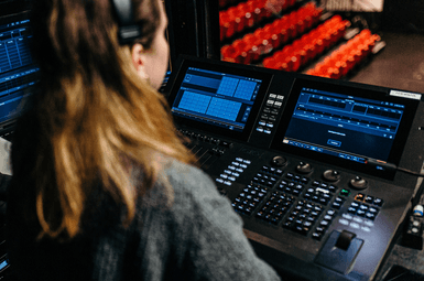 A lady sits wearing headphones, over a control technicians board.