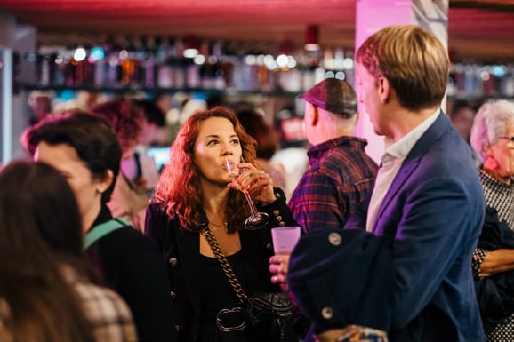 Audience members mingle in a crowded bar, lit in an atmospheric pink. In the centre is a lady sipping a glass of fizz, looking up at a man whose back is to the camera, listening. They look relaxed and casual.
