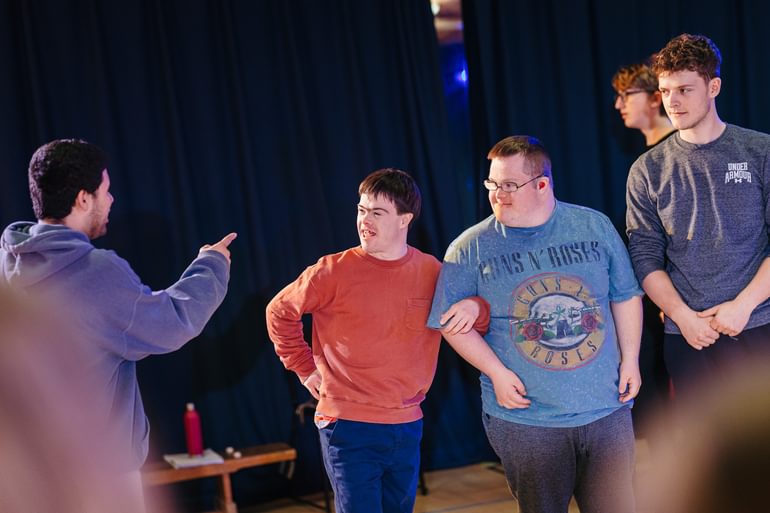 Four young people rehearse together with a dark curtain in the background. One stands with their back to the camera pointing at the others., who are looking at them with animated expressions. Another performer also looks on, with a more concerned and serious expression.