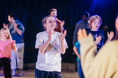 Young people perform together with a dark curtain behind them, lit in white and blue lighting. They are jumping and clapping, in the middle of a dance. The performer in the centre smiles enthusiastically and they all look like they're having a great time.
