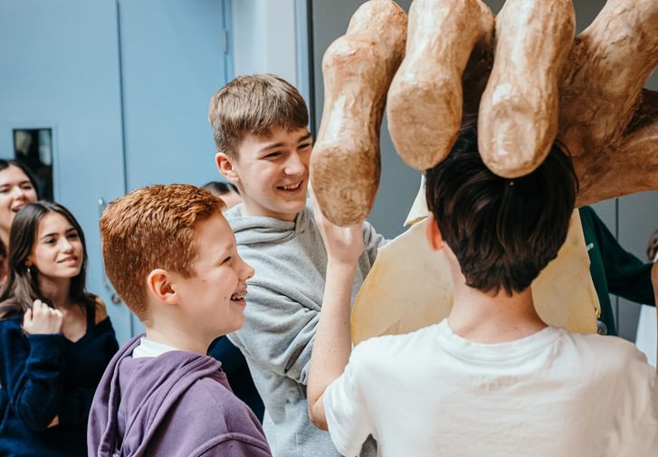 Three young performers hold up a huge oversized model hand. They are smiling and enjoying themselves. Other young performers look on from the background.