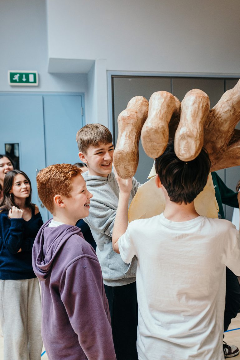 Three young performers hold up a huge oversized model hand. They are smiling and enjoying themselves. Other young performers look on from the background.