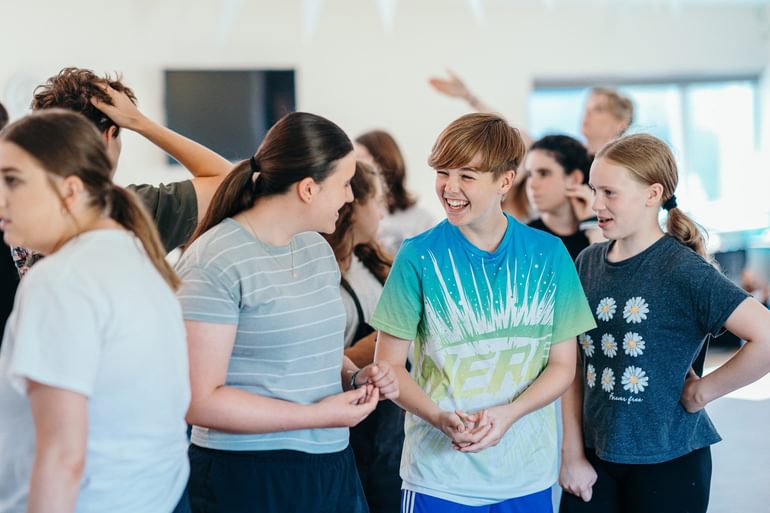A group of young performers stand together in a rehearsal room, talking and laughing together. They look relaxed and like they are having a really great time.