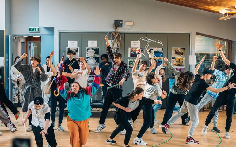 Lots of young performers rehearse together in a studio. They are all posing in dynamic positions with arms outstretched, facing out in all directions. There is an exciting, tense energy in the room. They wear colourful clothes and some hold props out in the air.