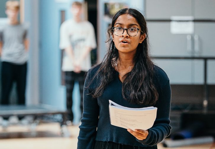 A young performer sings passionately in a rehearsal studio, holding a script.