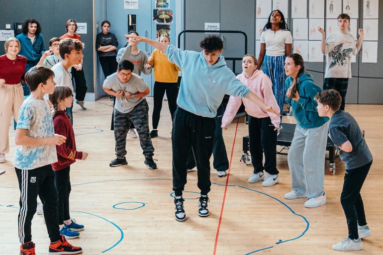 A group of young performers rehearse in a studio. The wooden floor has been marked out with different coloured tape so it looks a bit like a gymnasium. In the centre, a performer stands with arms outstretched as though balancing on a tightrope. The other performers gather around them in a semi-circle, looking on with curiosity and amazement. It looks fun and exciting.