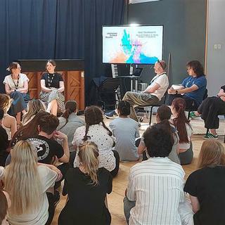 A group of Chichester College students sit on the floor infront of a panel of speakers and a TV screen.