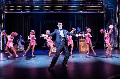 Charlie Stemp stands with arms outstretched on a stage. Girls in pink holding telephones are lines up behind him.