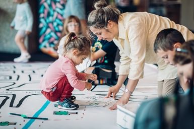 A small girl sits on a giant map. She is sticking a paper shape to it with a grown-up.
