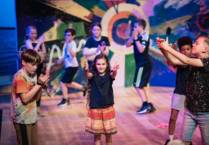 A girl is on a colourful stage with her arms outstretched. There are other children performing in the space around her.