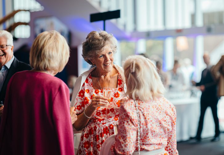 Three women chat animatedly to each other in the theatre foyer; the woman in the centre wears a party dress of orange flowers on white and carries a glass of champagne