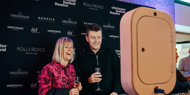 A man (in a suit jacket and shirt) and a woman (with a pink shirt and glass of fizz in hand) smile warmly as they pose for a picture