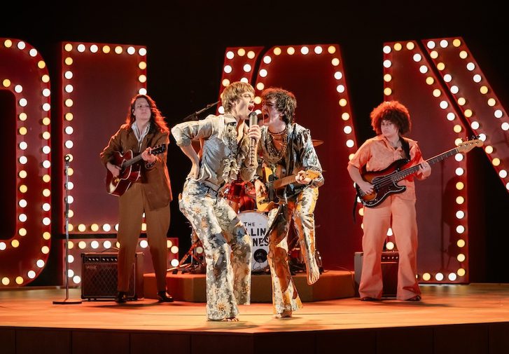 In the centre, two men in extravagant 1960s costumes - flowery flares and silvery satin shirts with ruffles - are giving an energetic performance. The man on the left is singing into a hand-held microphone with his right elbow bent and his hand on his hip. The musician on the right is also singing into his companion's microphone while playing a guitar. Behind them are two more guitarists and in the background are a number of large illuminated capital letters.