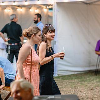 Two women in summer dresses are walking across the grass outside the Festival Theatre with drinks in their hand, enjoying the pre-show buzz.