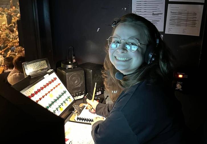 Zoe is sitting in a darkened sound booth with her face lit from below by the lights on the control panel. She has a beaming smile on her face as she looks towards the camera.