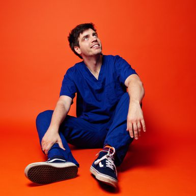 An image of Ed Patrick wearing blue medical scrubs and trainers, sitting looking up towards the ceiling. He is sitting on an orange backdrop.