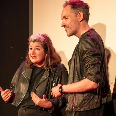 A photo of two people (Laura and Paul) on stage. Laura is shorter than Paul has shoulder length brown hair with a blonde quiff, she is wearing a black top and black leather jacket and making a gesture motioning towards herself. Paul stands next to her also dressed all in black.