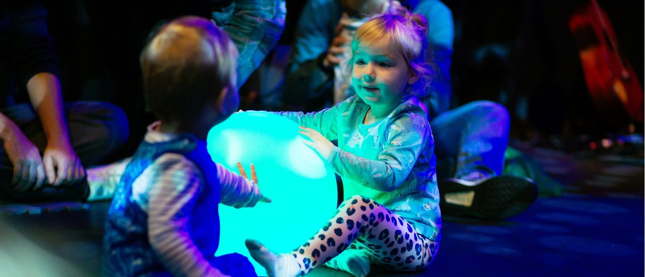 Two toddlers play around a large ball which is lit up with blue light from within.