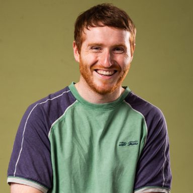 Ali Woods has short red hair and beard. He is smiling looking straight to camera. He is wearing a green t-shirt with blue short sleevs.