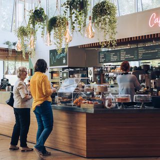 Two customers are waiting at the counter in our cafe which is loaded with delicious looking cakes. Behind the counter a staff member is making barista coffees.