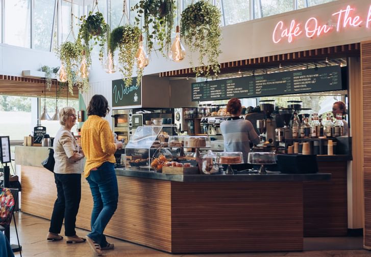 Two customers are waiting at the counter in our cafe which is loaded with delicious looking cakes. Behind the counter a staff member is making barista coffees.
