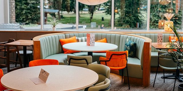 A view of the Minerva Bar, the main focus being a couple of tables, one of which is a booth with colourful cushions on the seats leading to the greenery of Oaklands park out of the window.