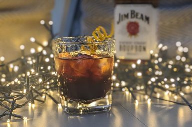 A Winter's night cocktail. A short glass filled with ice and dark colour liquid. Fairy lights sit behind and a bottle of Jim Beam behind.
