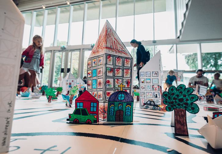 A close up of some hand-drawn 3D versions of Chichester landmarks in the middle of a giant paper map. The map sits in the middle of the theatre foyer with children running around it and cutting out other landmarks.