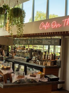 A view of a cafe counter laden with cakes and baked goods. A pink neon sign reads 'Cafe on the Park' and plants hang above the counter.