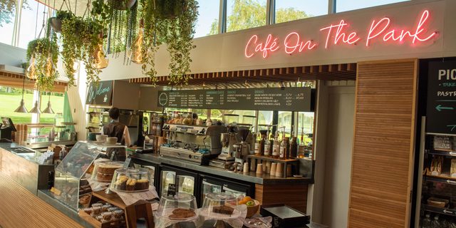 A view of a cafe counter laden with cakes and baked goods. A pink neon sign reads 'Cafe on the Park' and plants hang above the counter.