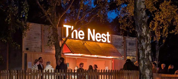 A nighttime scene, with an illuminated sign 'The Nest' above a wooden building between trees. People are gathered in front of a bar lit with warm-tones, on a raised terrace behind a wooden picket fence.