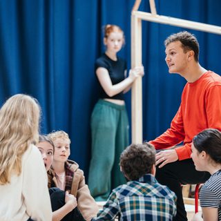 A group of young performers are sat on the floor talking to one another. A boy in a red top is sat on a chair looking down at them. Behind, two girls are holding a large wooden frame.