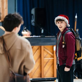A young performer in a burgundy jacket and red Santa hat is talking to someone whose back is facing the camera. They have a large backpack with a torch sticking out of the top.