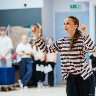 A young girl in a pink and black striped top is stood with her arms reaching in front of her. She looks excited. Other performers are stood watching in the background.
