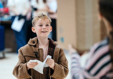 A young boy in a brown trench coat smiles gently. He is holding a folded piece of paper in his hands.